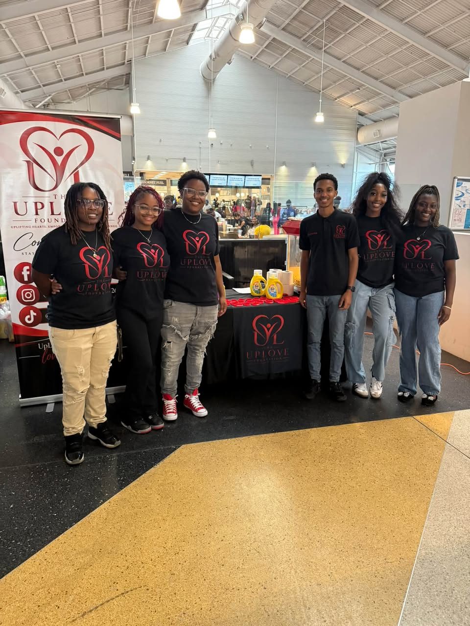 Six UpLove Foundation volunteers standing beside their branded table and pull-up banner in the university dining hall