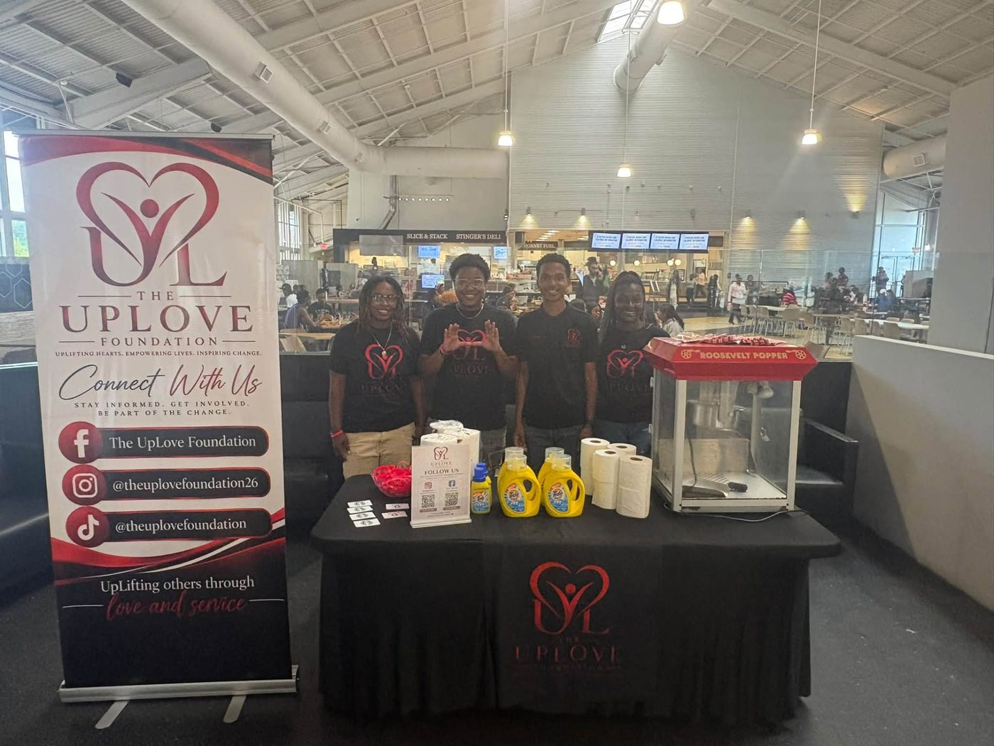 Four UpLove Foundation volunteers behind their display table stocked with laundry detergent, paper towels and a popcorn machine